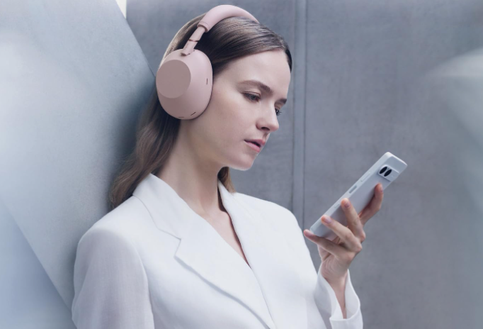 Woman wearing pink Sony WH-1000XM6 over-ear headphones while holding a phone