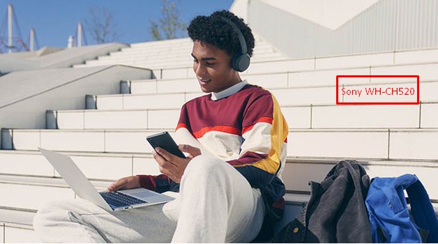 Man wearing black Sony WH-CH520 headphones sitting on outdoor steps with a phone and laptop
