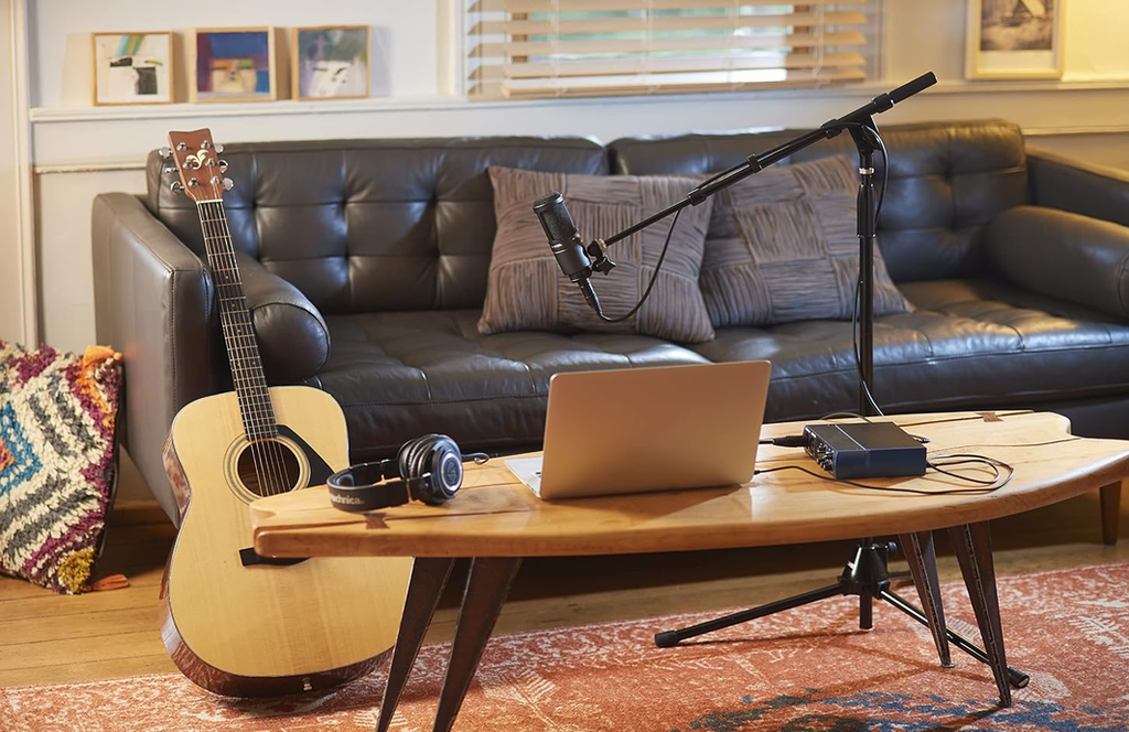 Wide view of a home recording setup with the Audio-Technica AT2020 on a boom arm above a table with a laptop and guitar beside a sofa